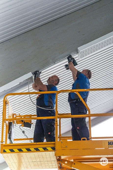 Elektriker von Marquardt & Streck montieren Leuchten unter dem Hallendach einer Turnhalle